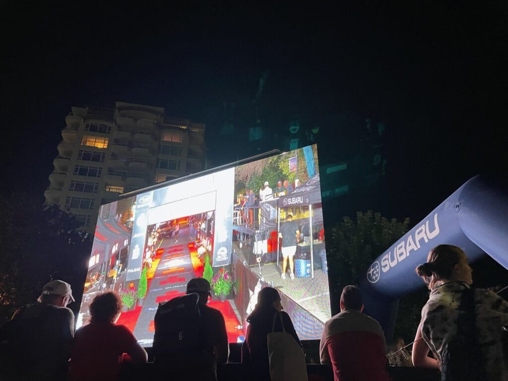 A crowd of spectators gathers in front of a jumbotron screen positioned near the finish line of a race. Audience Engagement.